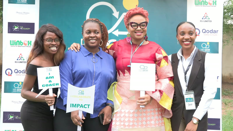 Four members smiling in front of the HR GEM Coach Network backdrop, holding "I am a coach" and "Impact" placards