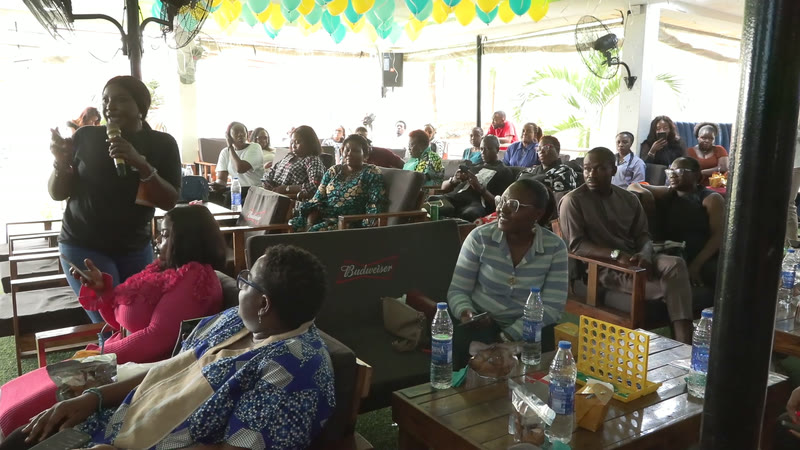 A large audience seated at tables under a canopy, listening to a speaker