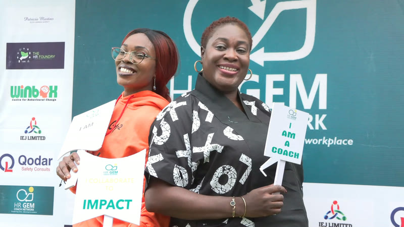 Two smiling members holding "Impact" and "I am a coach" placards at the HR GEM backdrop